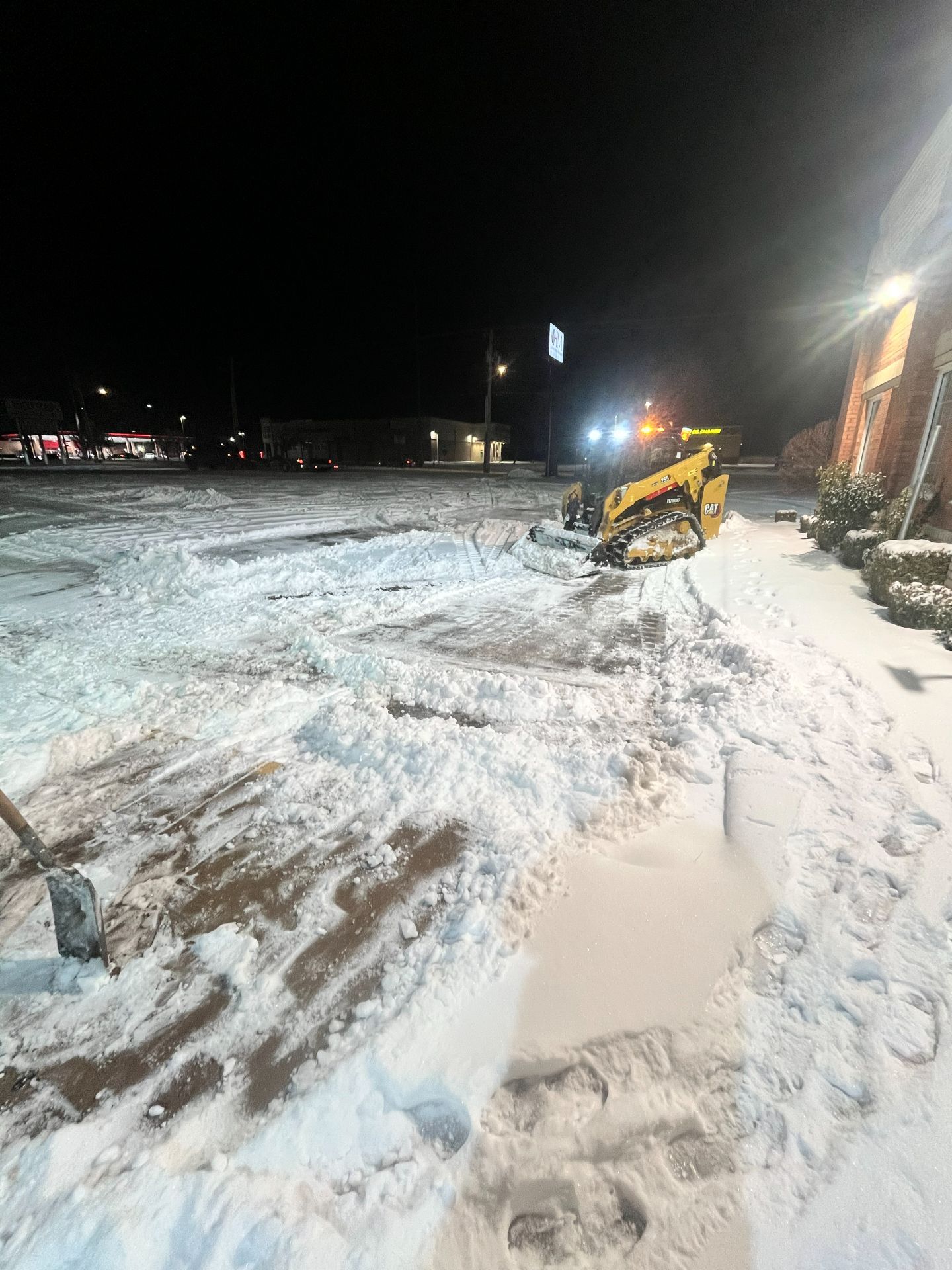 A yellow skid steer clears snow from a parking lot at night in front of a building.