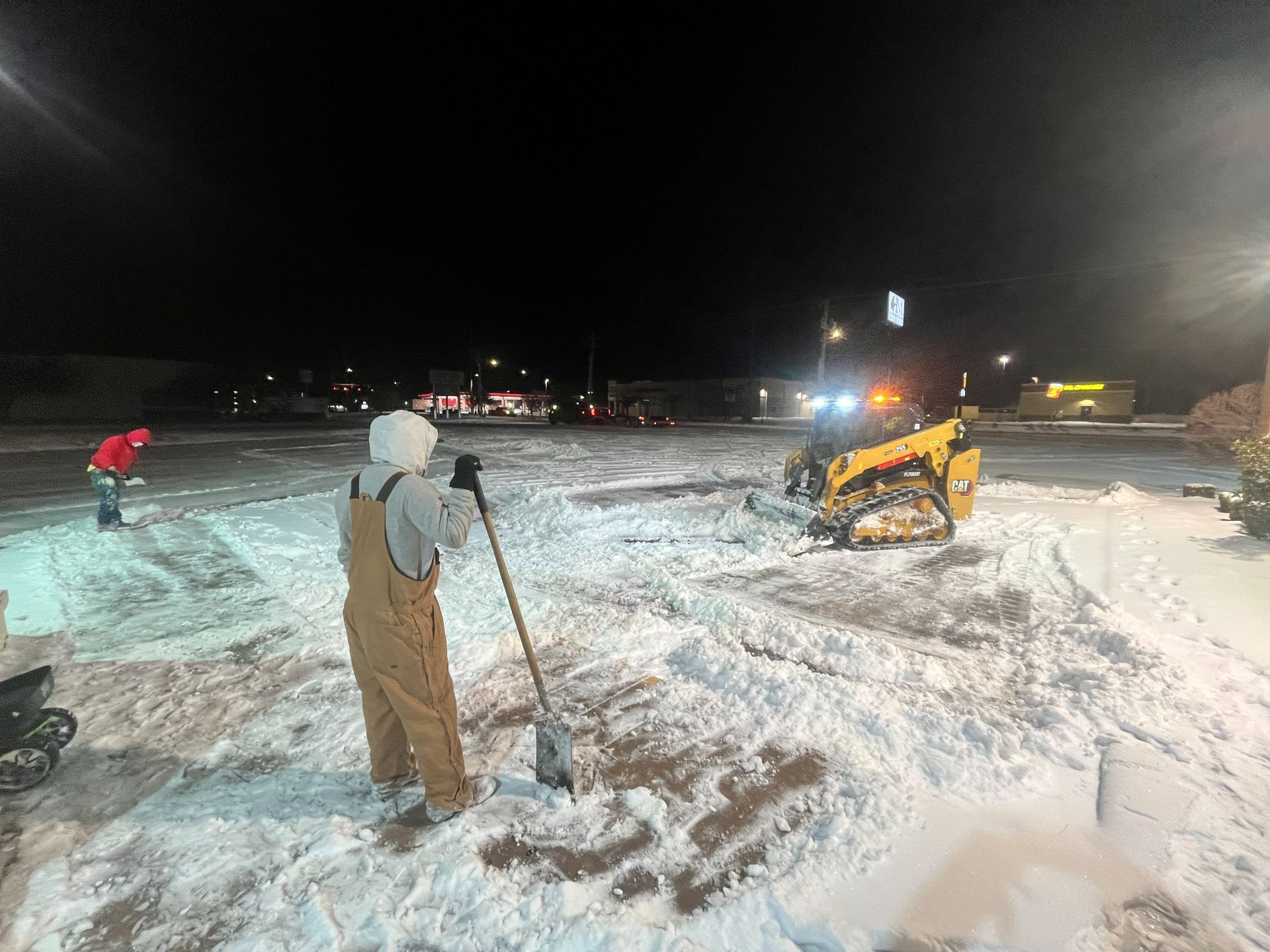Workers in a parking lot at night use shovels and a yellow skid-steer loader to clear snow.