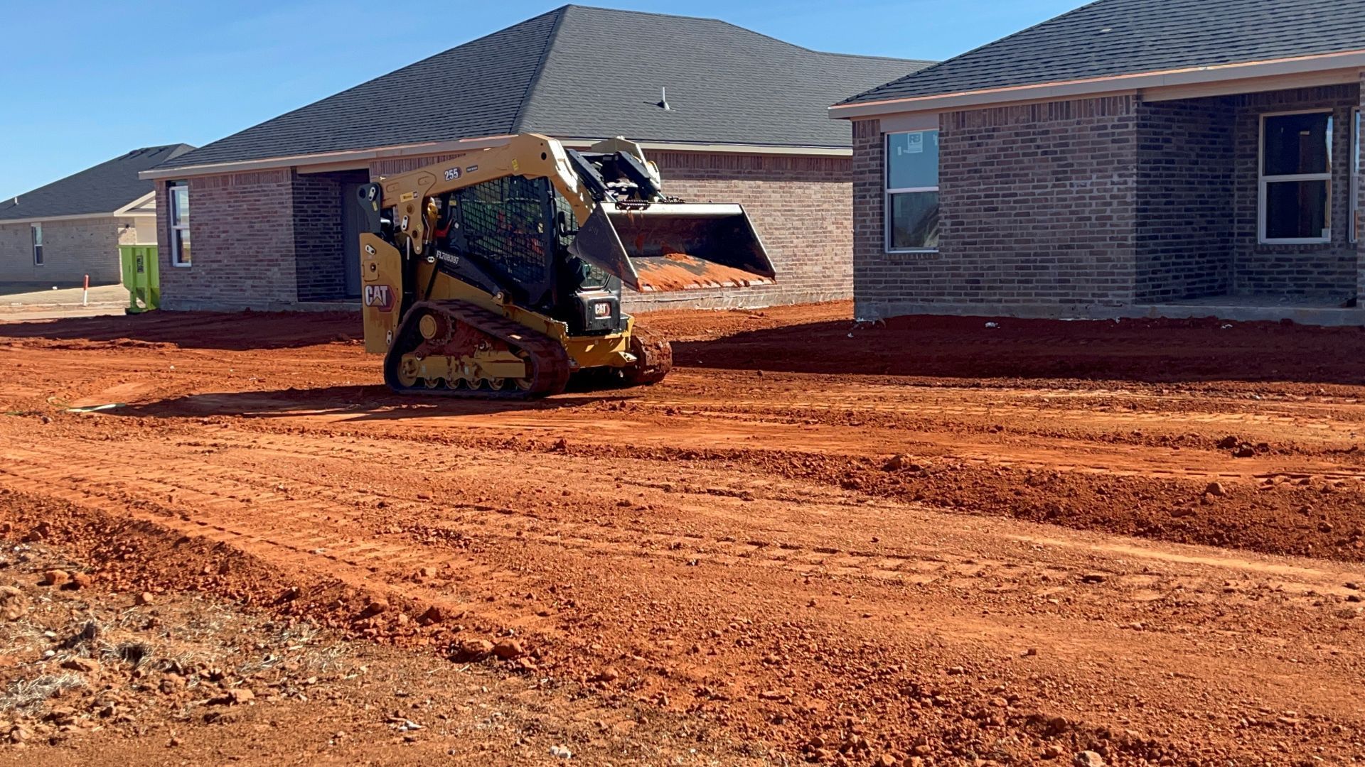 A yellow skid steer loader moves red soil at a construction site between two new brick houses under a clear blue sky.