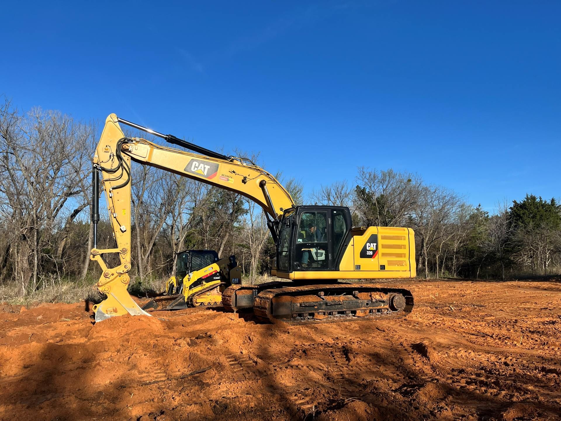 A yellow Caterpillar excavator sits on a patch of red dirt against a bright blue sky with trees in the background.