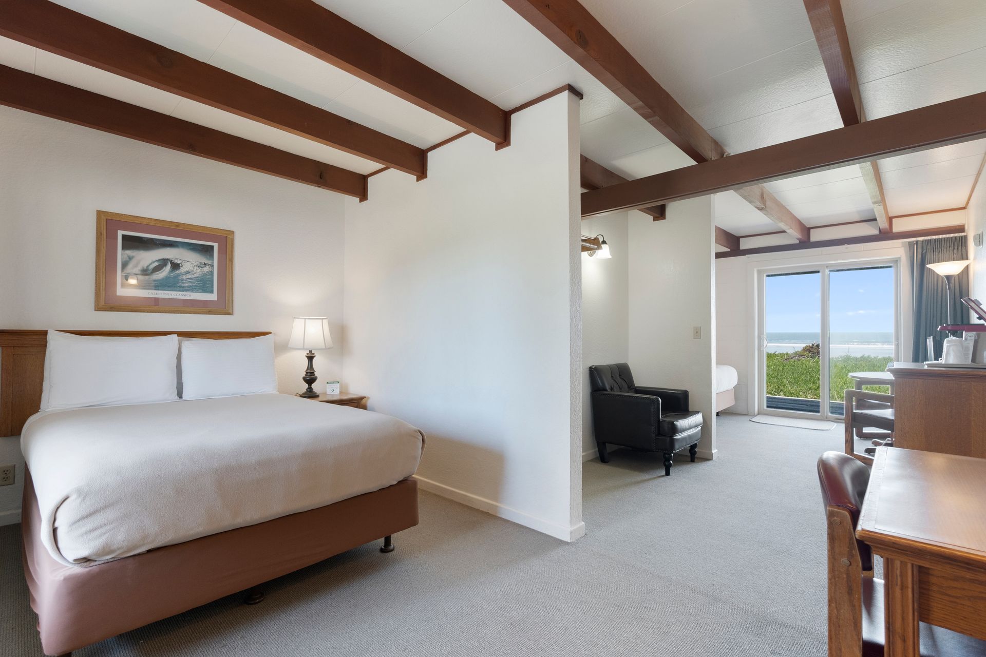 Hotel room with a bed, armchair, desk, and ocean view through a sliding glass door.