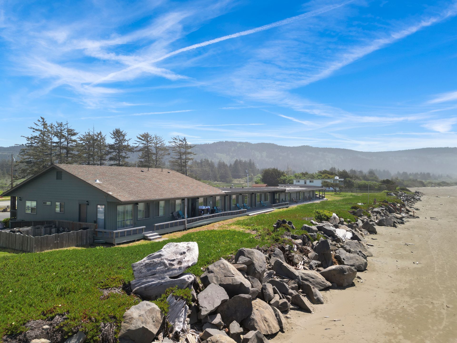 Beachfront motel with green exterior under a blue sky, next to a rock wall and sandy beach.