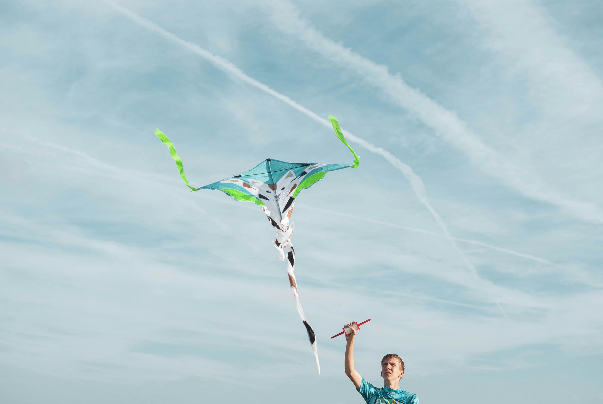 Person flying a colorful kite against a blue sky with contrails.