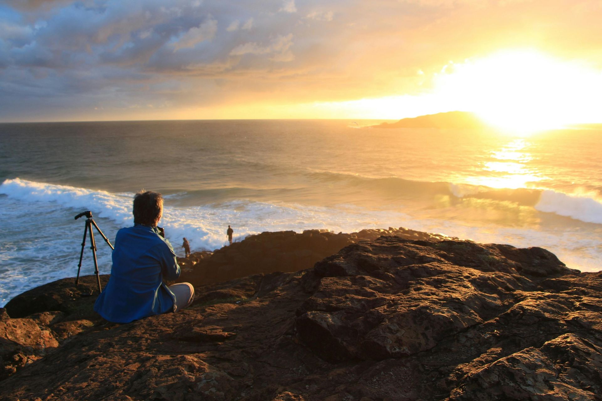 Photographer at sunrise overlooking ocean waves, golden sky.