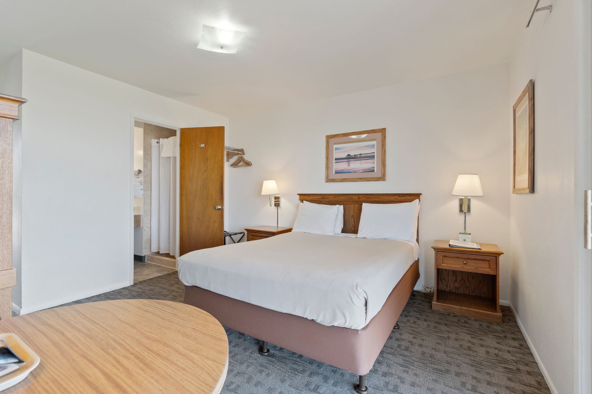 Bedroom with a bed, nightstands, a bathroom door, and a table. The room is brightly lit and has a neutral color scheme.