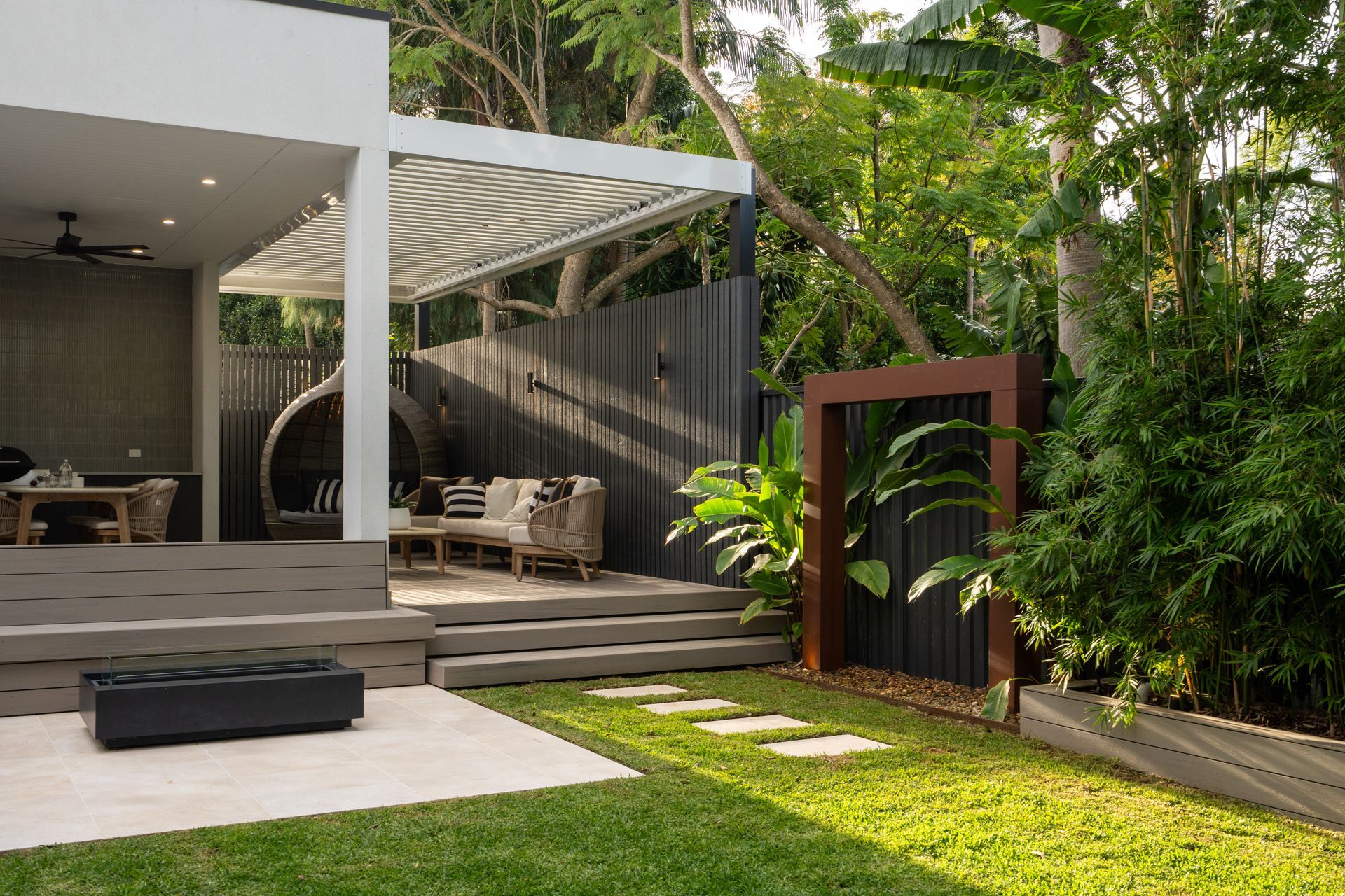 A backyard with an outdoor entertaining area, patio, privacy screenng and waterfeature surrounded by trees.