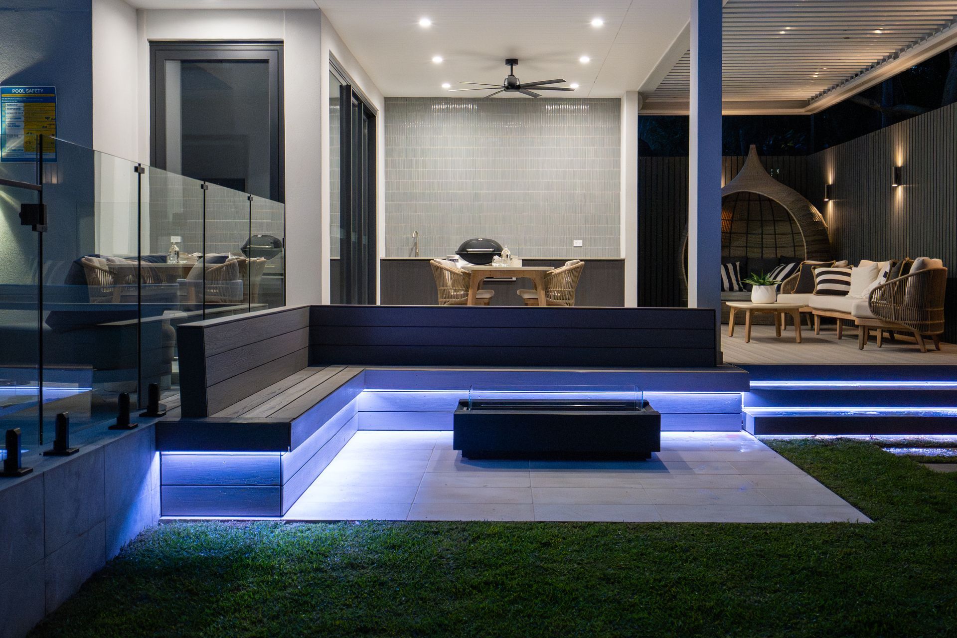 A modern backyard with a couch , outdoor kitchen, dining table, firepit and lighting.