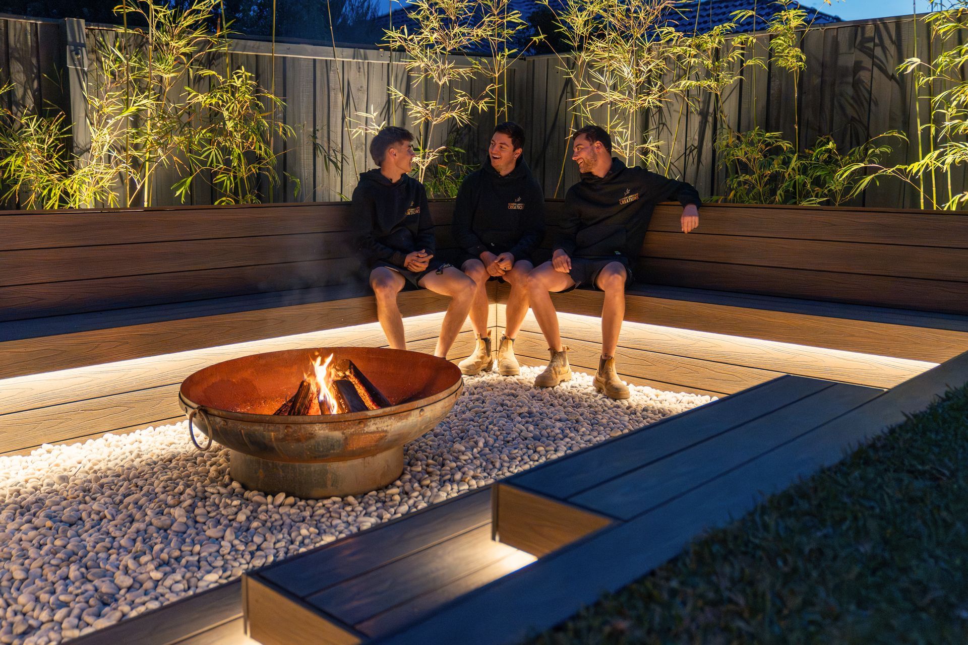 Three men are sitting on a bench next to a fire pit.