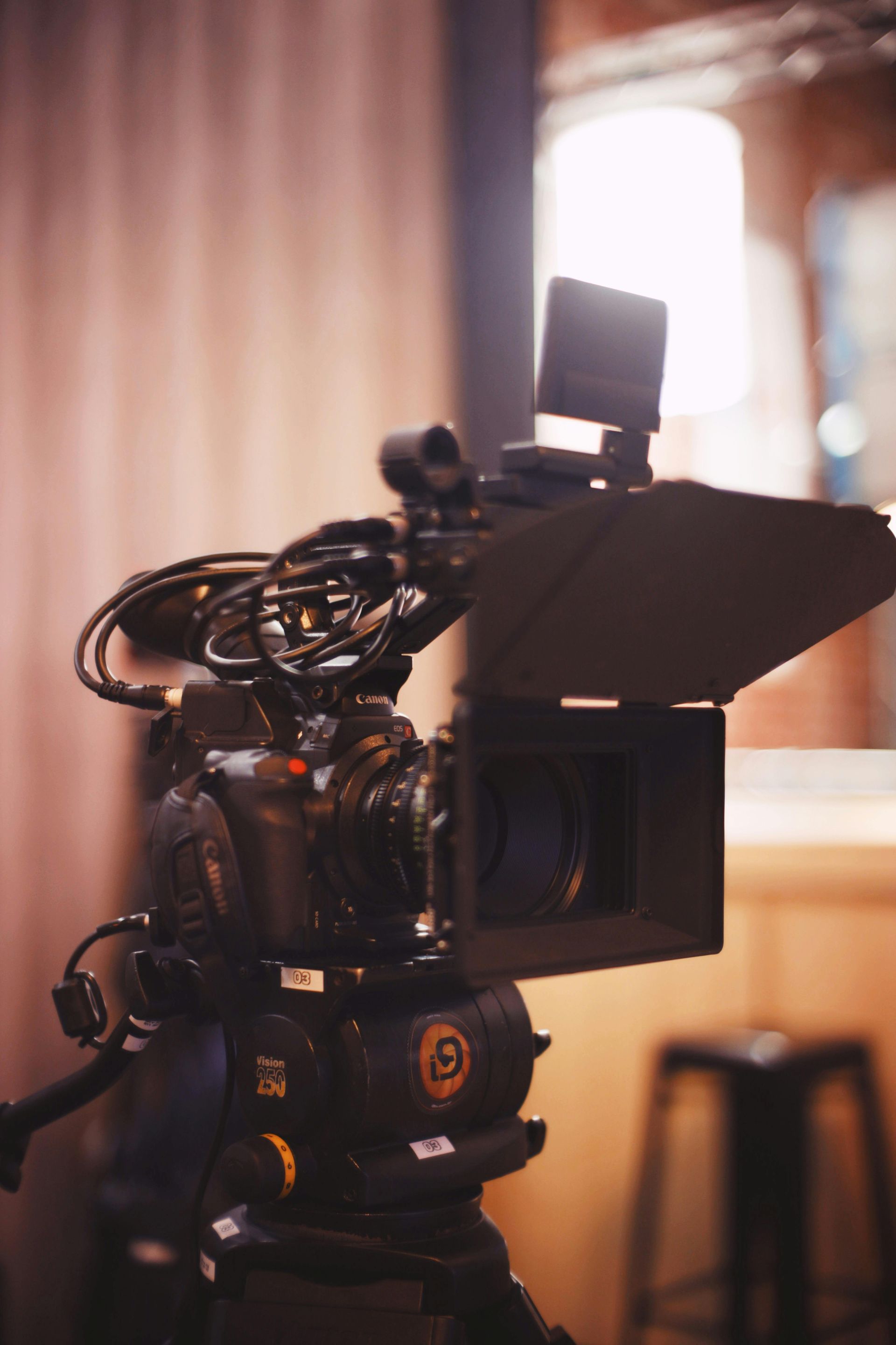 A professional video camera with teleprompter mounted on a tripod indoors, filming a blurred background.