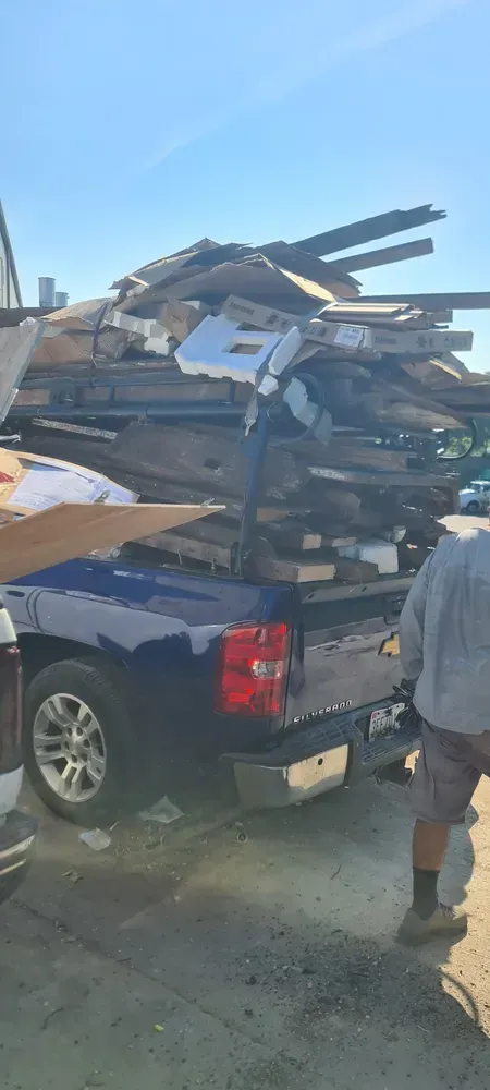 A blue pickup truck filled with wood, a person standing nearby on a concrete surface under a clear sky.