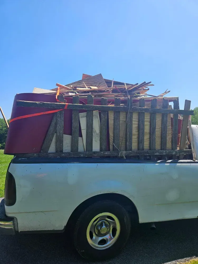 White pickup truck bed filled with wood, a red couch piece, and debris, secured with straps, under a blue sky.
