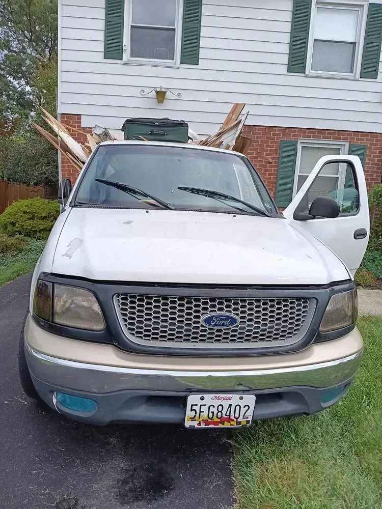 White Ford pickup truck with a tree branch on top and the door open, parked in front of a house.
