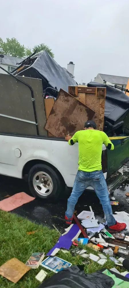 Man loads junk into a truck on a wet day; debris on lawn; house in background.