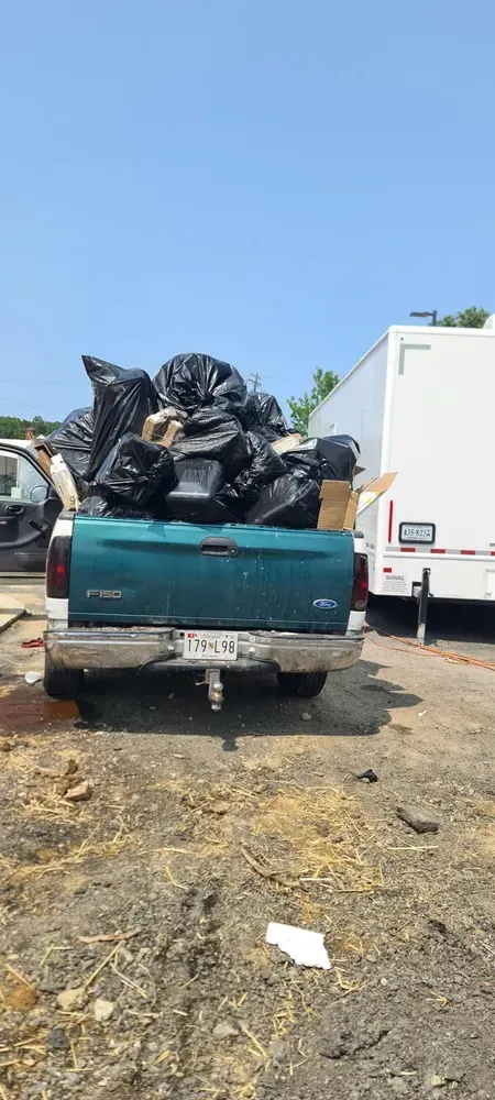 A teal truck bed overflowing with black trash bags, parked outside, with a clear blue sky above.