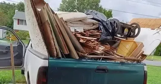 Green pickup truck bed overflowing with debris, including wood, a mattress, and a tire.