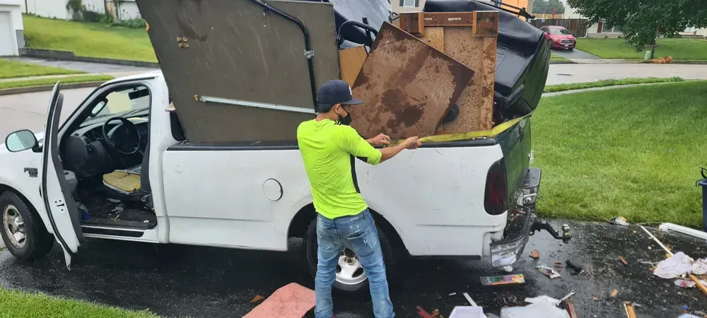 Man in green shirt struggles to secure items in white pickup truck on a residential driveway.