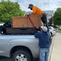 Two men loading furniture onto a silver pickup truck, one standing on the truck bed securing the items with a strap.