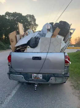 A silver pickup truck bed is overflowing with construction debris on a roadside.