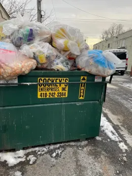 Green dumpster overflowing with trash bags; parked on snowy ground.