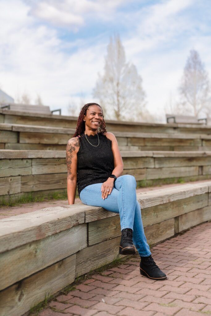 Woman sits smiling on wooden steps, wearing black top, jeans, boots. Outdoors, cloudy sky.