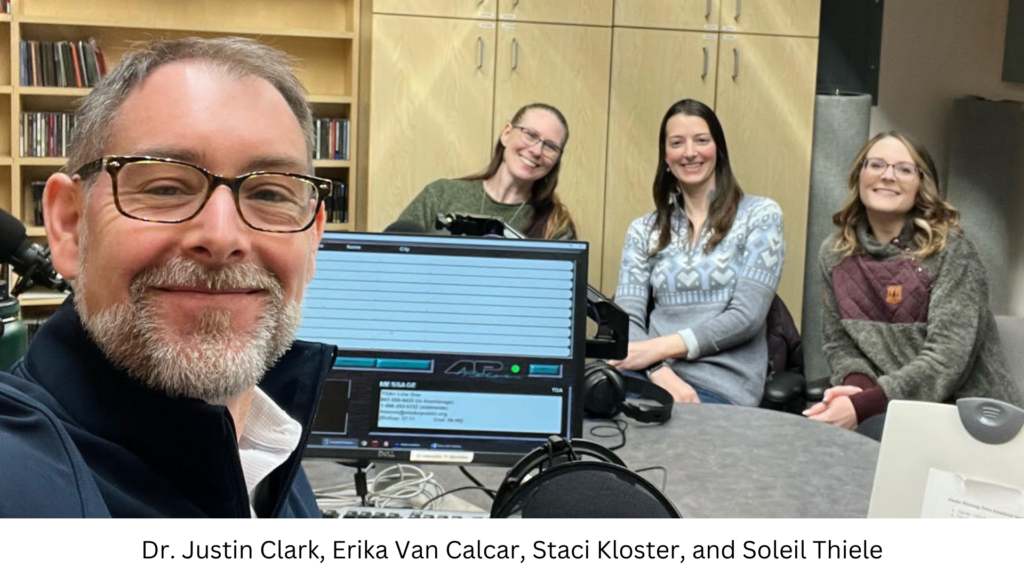 Dr. Justin Clark with three women at a desk with audio recording equipment. They are smiling in a studio.