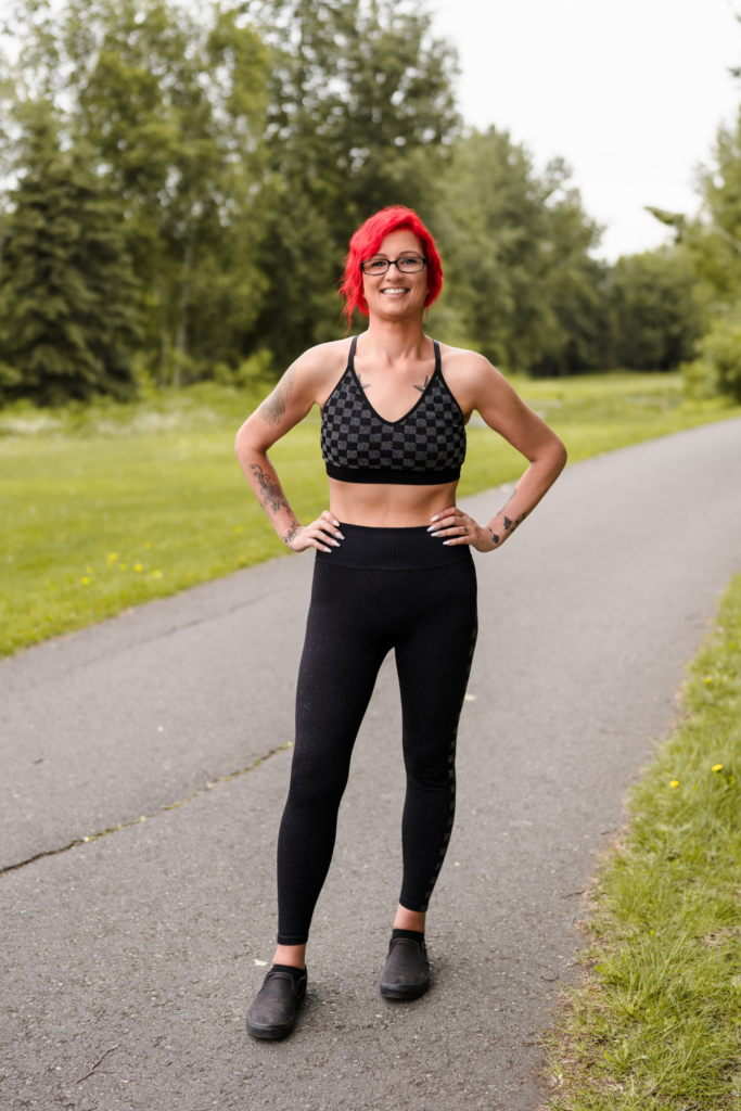 Woman with red hair in workout clothes smiles on a paved path outdoors.