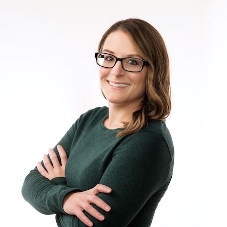 Woman with glasses and arms crossed, smiling, wearing a green sweater against a white backdrop.