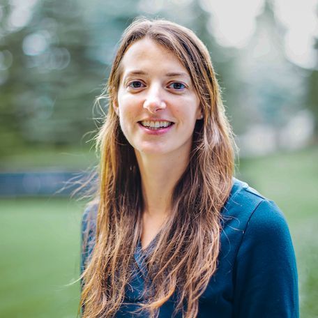 Woman with long brown hair smiles outdoors in front of blurred trees. She wears a blue top.