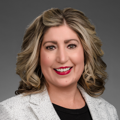 Woman with blonde hair, wearing a black top and a white blazer, smiles at the camera.