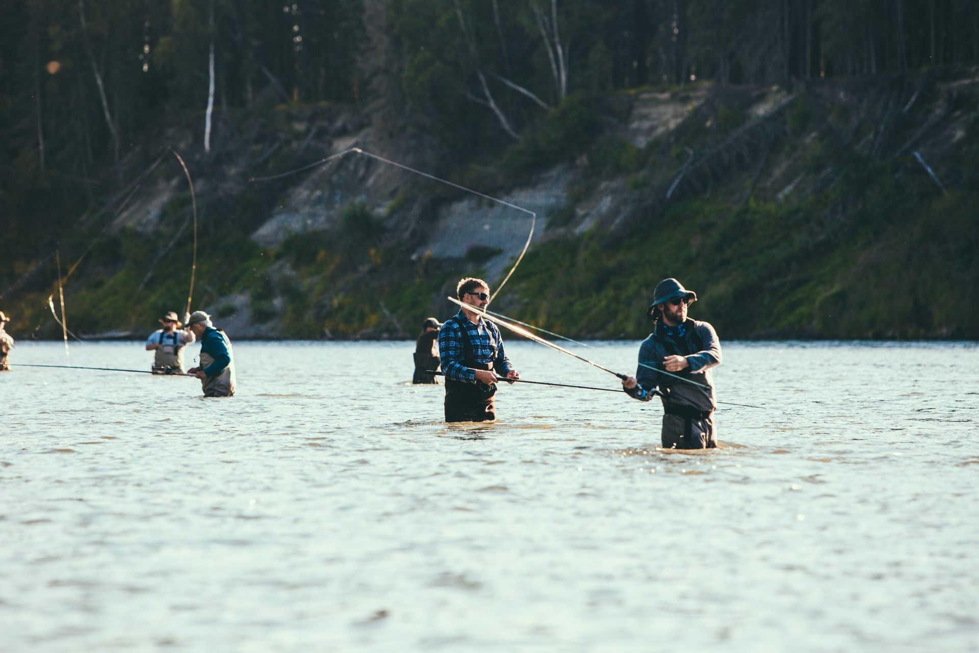 People fly fishing in a river, wearing waders.  Green trees line the shore.