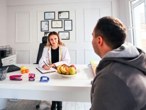 Dietitian consults with a patient at a desk with fruit, office supplies, and framed certifications.