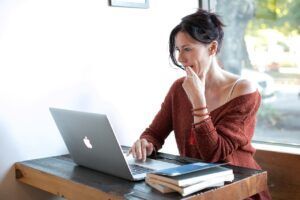 Woman sitting at a wooden table with laptop, thoughtfully touching chin, near window.