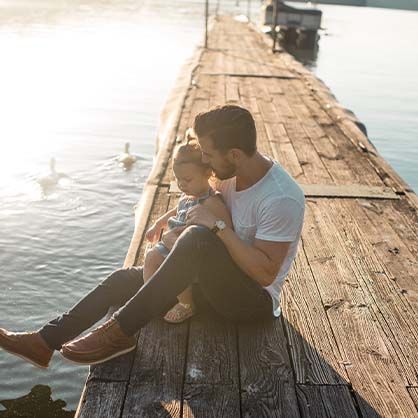 Man and child sitting on wooden dock, looking at water with swans.