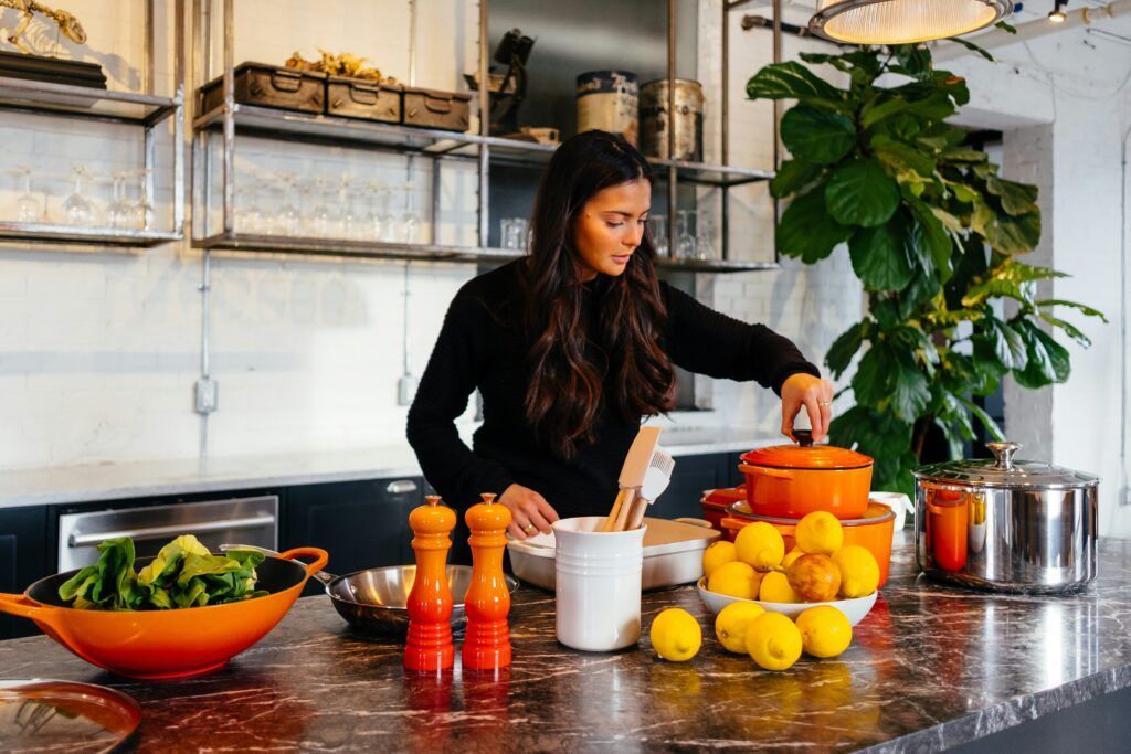Woman in a kitchen reaching for a pot. Brightly lit with lemons and vegetables on a countertop.