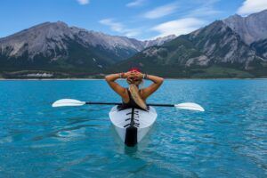 Woman in kayak on turquoise water, looking at mountains with hands on head.