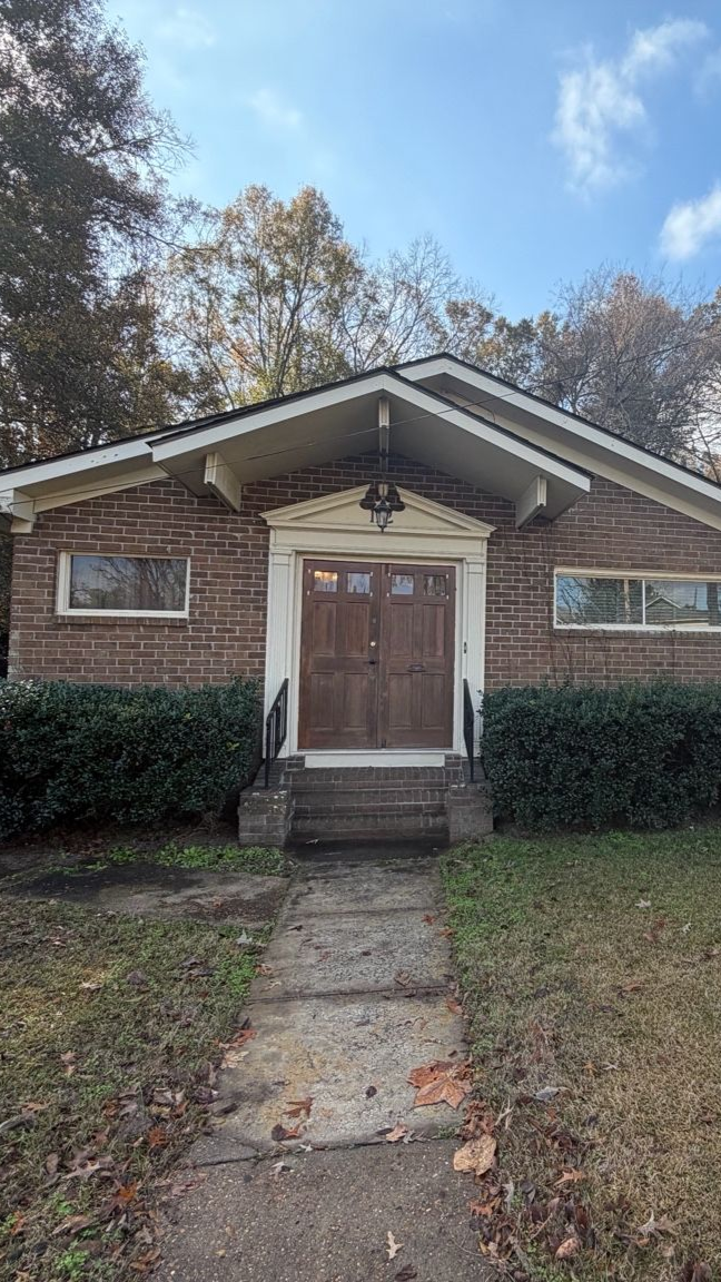 Brick building with a brown door, topped by a decorative roof. Bushes line the front.