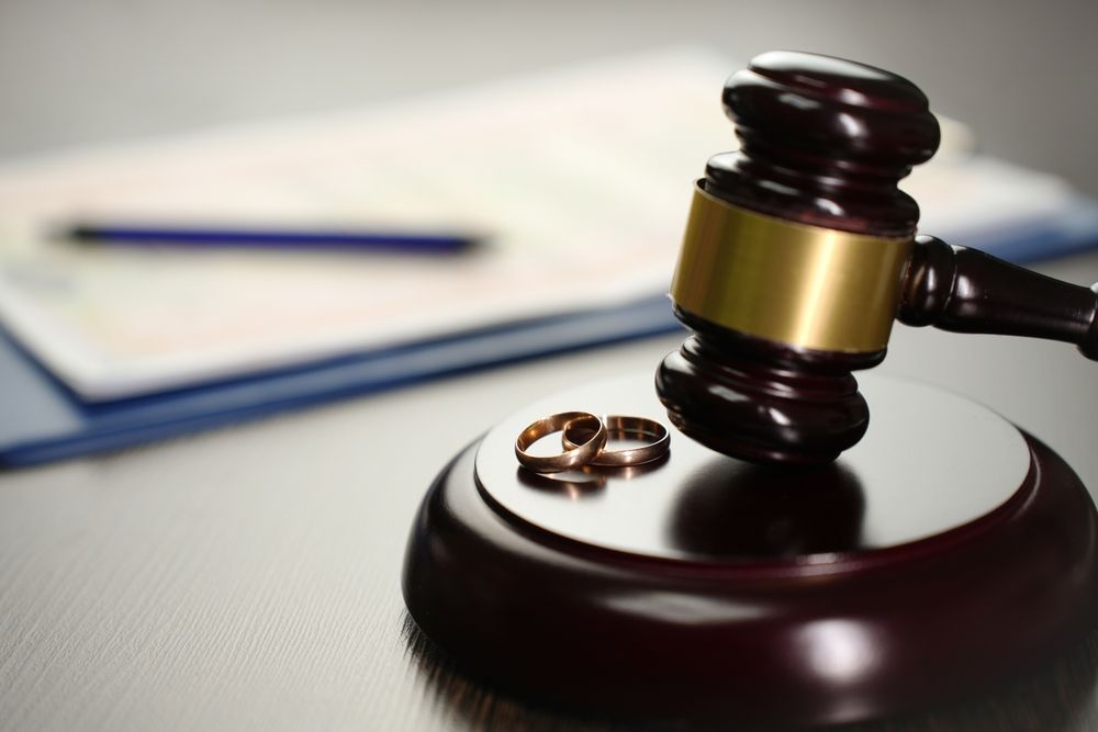 Gavel resting on a wooden block, with two wedding rings beside it. Blurred document and pen in the background.