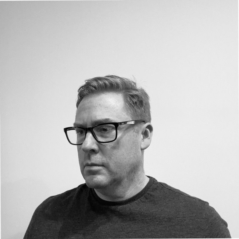 A man wearing glasses and a black shirt is standing in front of a white wall.