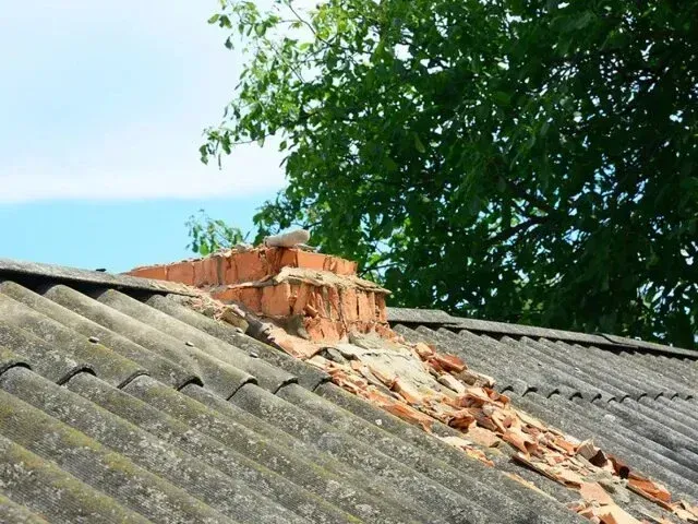 Chimney-Sweep-Portland - Damaged brick chimney on a slate roof with green trees and blue sky in the background.