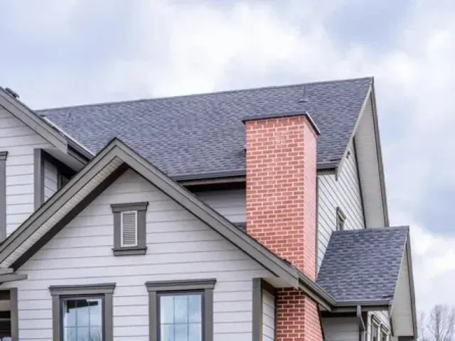 Chimney-Sweep-Portland- Close- up view of a modern house with a grey roof, white siding, and a prominent red brick chimney under a cloudy sky.