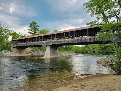Chimney-Sweep-Portland-Long wooden covered bridge spans a flowing river under a dramatic cloudy sky, surrounded by lush green trees.