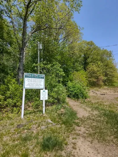 Chimney-Sweep-Portland-A sign for the Roscoe & Elva Lowell Preserve stands beside a dirt path leading into a wooded area with a clear blue sky above.