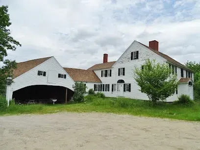 Chimney-Sweep-Portland-White colonial-style house with black shutters and a gambrel roof, surrounded by lush greenery. A covered walkway leads to a barn. Overcast sky. Serene atmosphere.