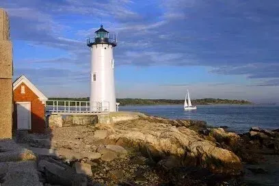 Chimney-Sweep-Portland-White lighthouse and red keeper's house stand on a rocky shore with a sailboat in the distance under a blue, cloudy sky.