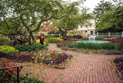 Chimney-Sweep-Portland-A woman stands in a lush garden with a fountain and a brick pathway in front of a white-fenced building.