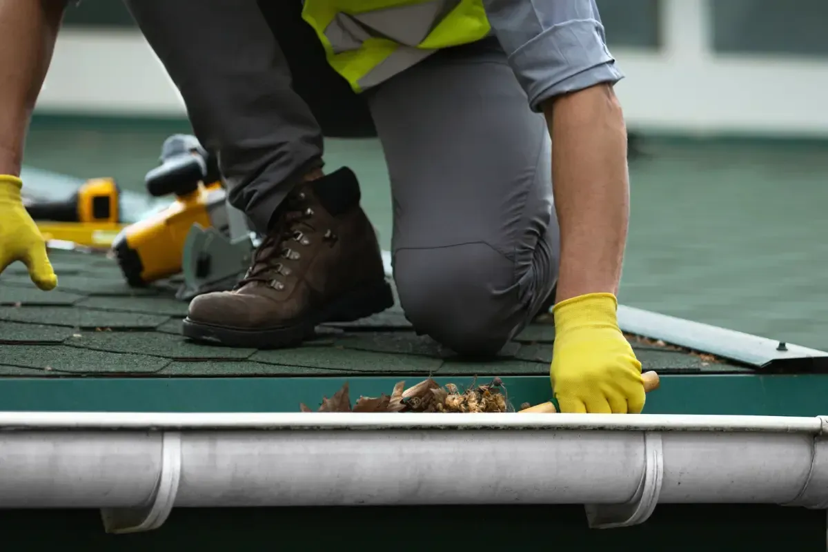 Person in work boots and gloves cleaning a gutter on a rooftop.