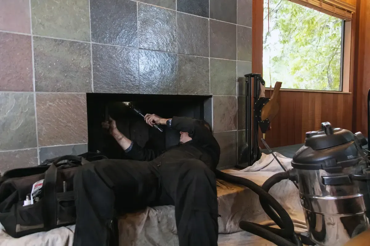 A person cleaning a fireplace, with a vacuum, tools, and a window in the background.