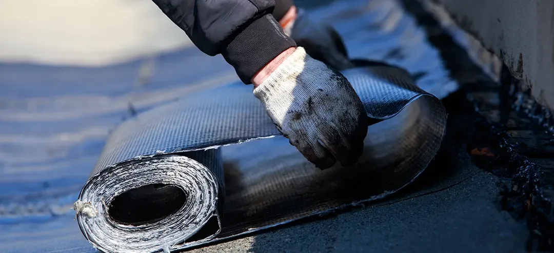A person is rolling a roll of roofing material on a roof.