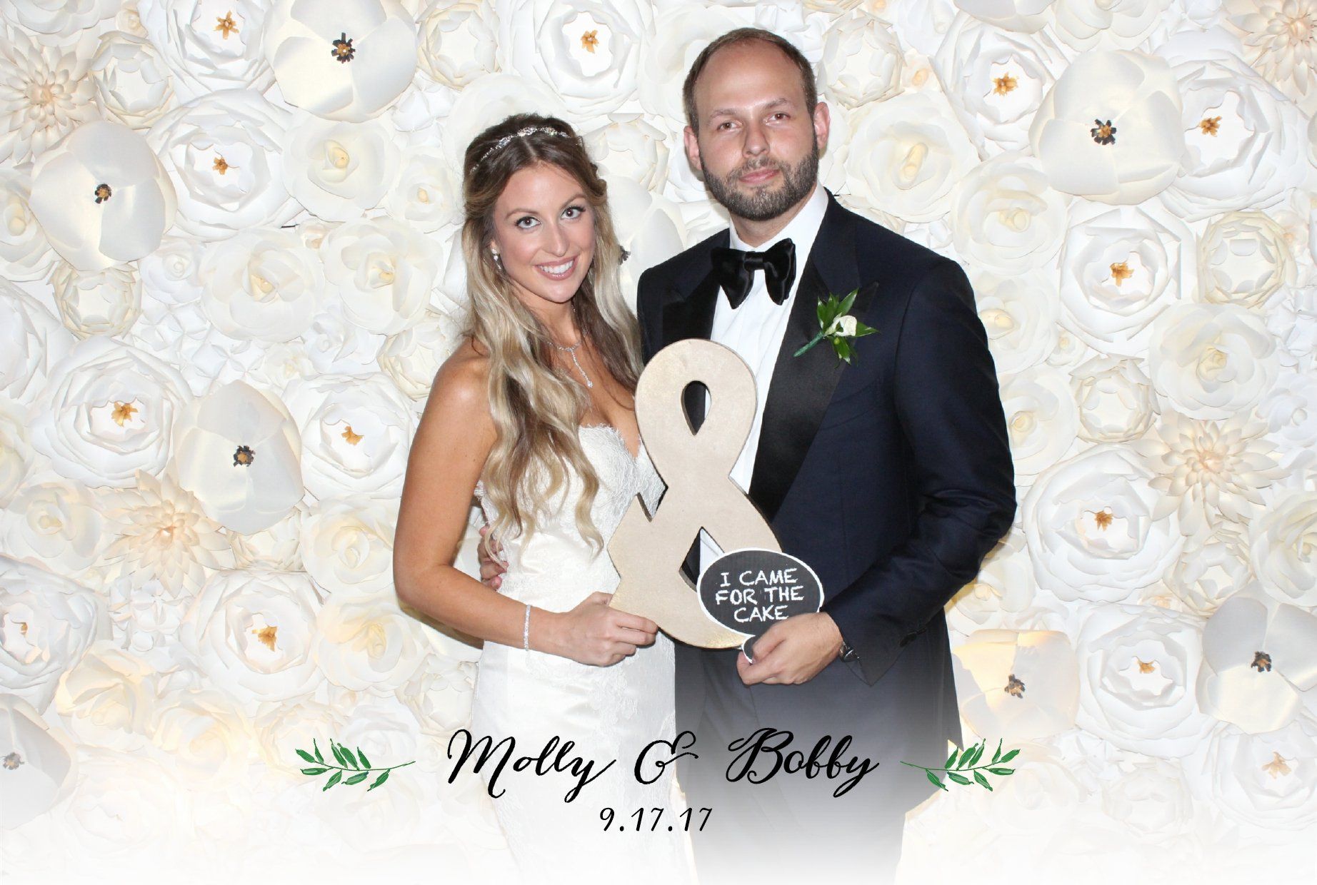 A bride and groom are posing for a picture in front of a wall of paper flowers.