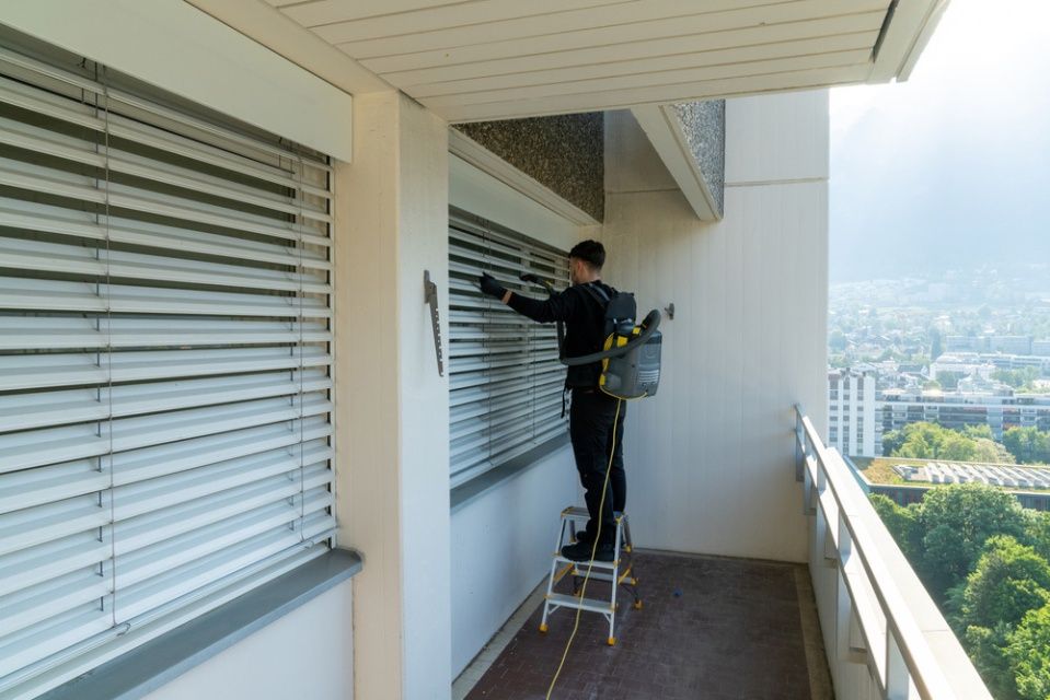 A Man Is Standing On A Ladder Cleaning Blinds On A Balcony — NJP Services In Coniston, NSW